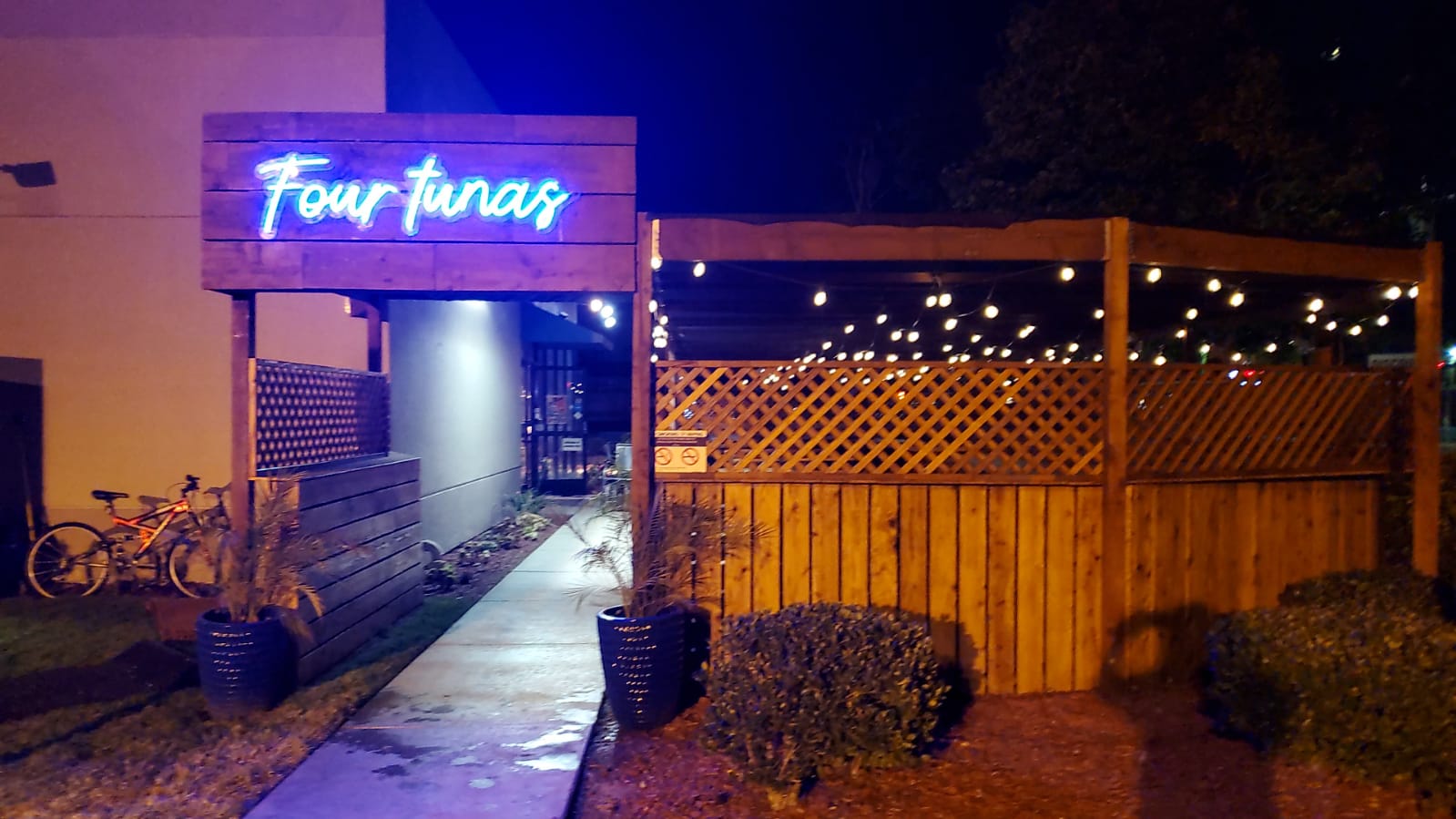 Outdoor dining area with a wooden fence, string lights, and a neon sign reading "Four Tunas" at night.
