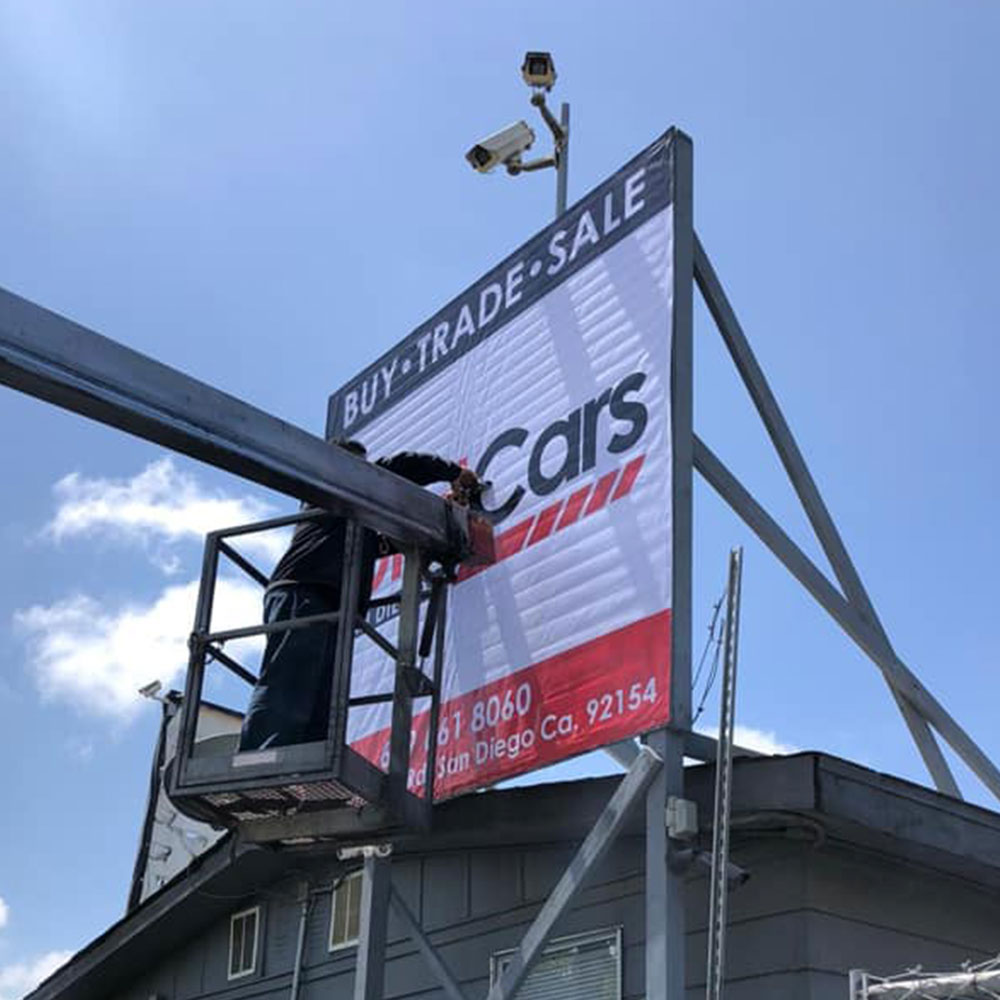 Worker on lift installing a large sign for a car dealership against a clear blue sky.