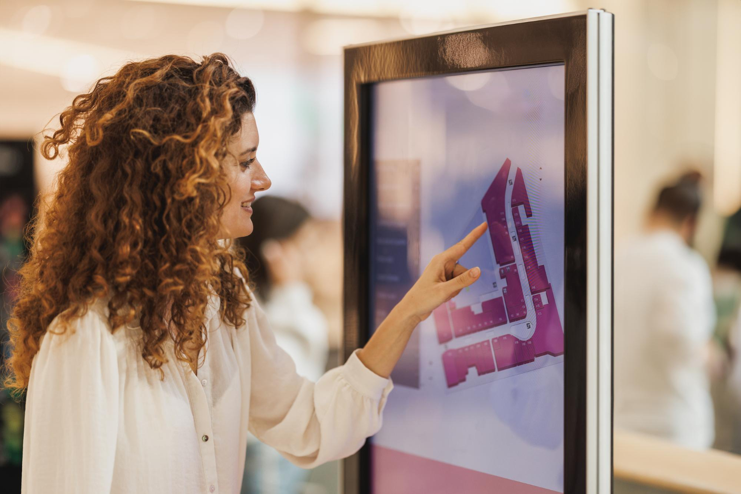 Woman using a mall directory touchscreen, pointing at a floor plan with a smile.