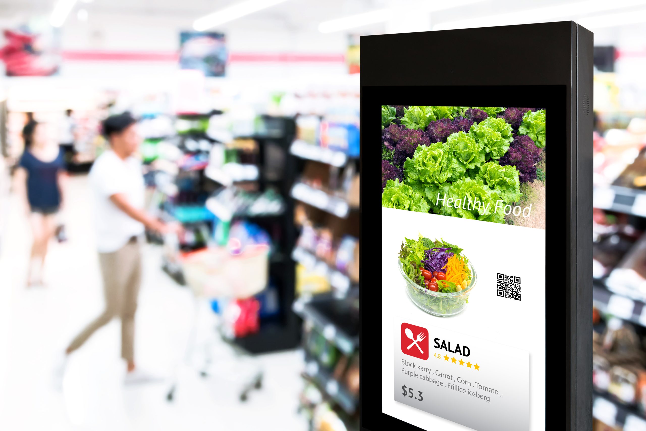 Digital display in a grocery store shows salad advertisement; shopper with a cart in the background.