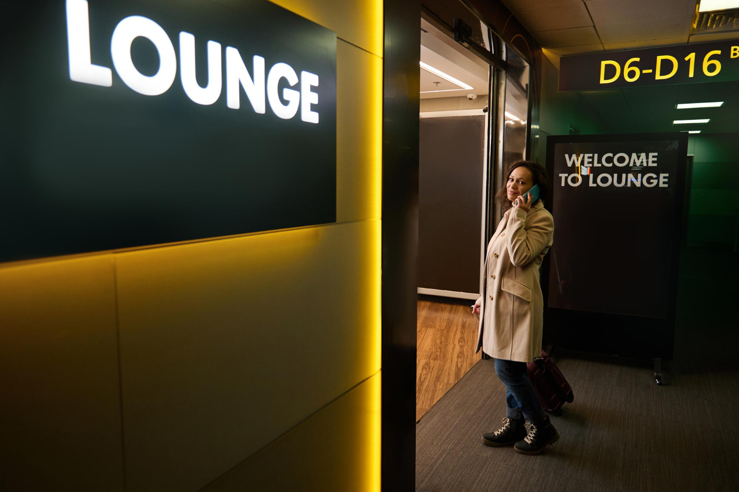 7800 Woman on phone with suitcase stands near airport lounge entrance with bright "LOUNGE" sign.